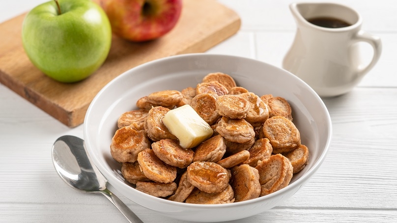 Apple cinnamon pancake cereal in a bowl next to apples and syrup.