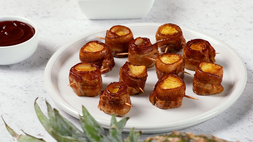 Bacon Wrapped Pineapple on white serving plate, dish of dipping sauce, white table