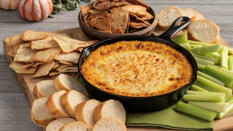 Baked Pumpkin Cheese dip in cast iron pan on top of wooden board with pita chips, sliced bread and celery sticks