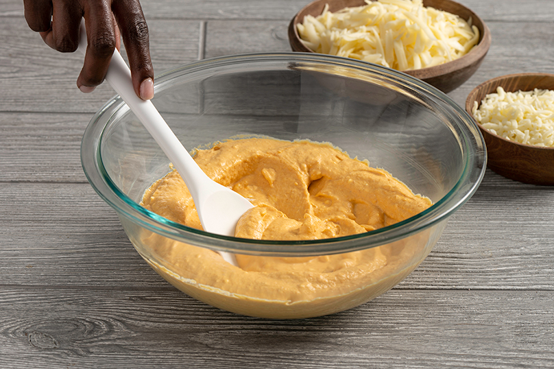 Hand mixing cream cheese and pumpkin in a medium bowl, serving dish of shredded cheese in background