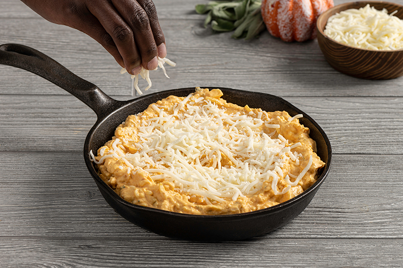 Hand sprinkling shredded cheese on top of pumpkin and cream cheese mixture in cast iron skillet