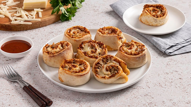 BBQ Chicken Pinwheels on white plate, cutting board with cheese and parsley in background, serving dish of barbecue sauce, fork, blue napkin, white stone counter background