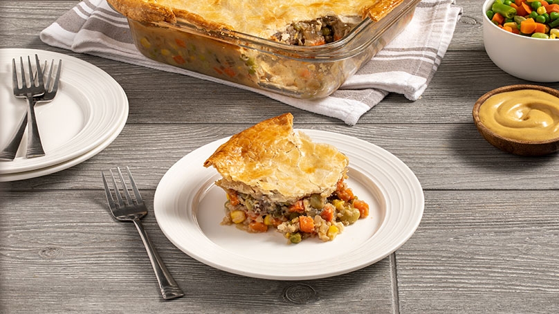 Beef and Beer pie on white plate in front of dish with rest of pie, serving dish of mixed vegetables and dijon mustard, fork and plates, gray wood table background