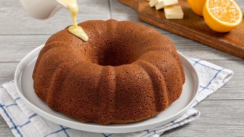 Brown Butter Bundt Cake on white serving plate, measuring cup pouring orange glaze onto bundt cake, cutting board in background with fresh halved oranges and butter chunks, blue checked napkin, gray wood table background