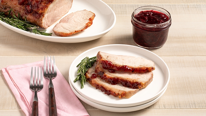 Cherry glazed pork lion on white plate with fresh rosemary, serving dish of entire cherry glazed pork loin in background next to jar of cherry preserves, pink napkin with two forks on top, light wood background