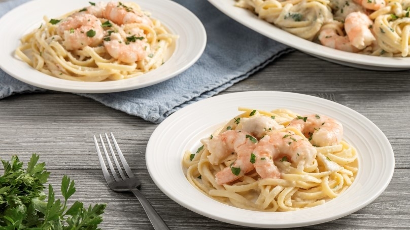 two white bowls of Creamy Garlic Seafood Pasta with fresh cilantro, blue napkin, fork, large plate of seafood pasta in background