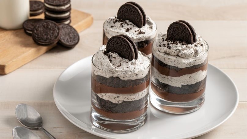 Five-ingredient cookies and cream parfaits on plate, chocolate sandwich cookies and milk on cutting board in background, spoons, wooden table