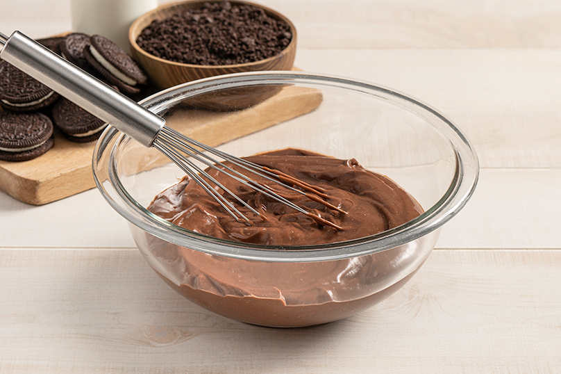 Clear mixing bowl with whisk mixing pudding mix and milk, chocolate creme sandwich cookies in background, wood table background