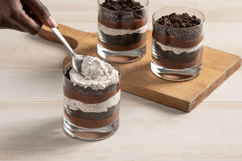 Hand layering whipped topping on top of cookie crumbs in glass to create cookies and cream parfait, two parfaits in background on wooden cutting board, light wood table