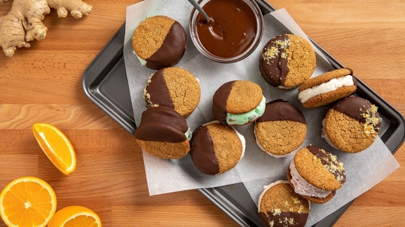 Ginger Cookie Ice Cream Sandwiches on baking sheet lined with wax paper, oranges, wood table