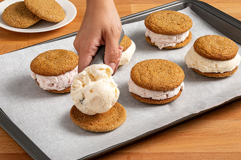 Hand scooping the ice cream onto a cookie on a lined baking sheet