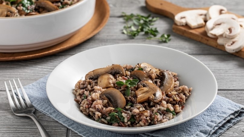 Hearty Mushroom Wild Rice with Spinach in white bowl on blue napkin, fork, serving bowl, cutting board with ingredients