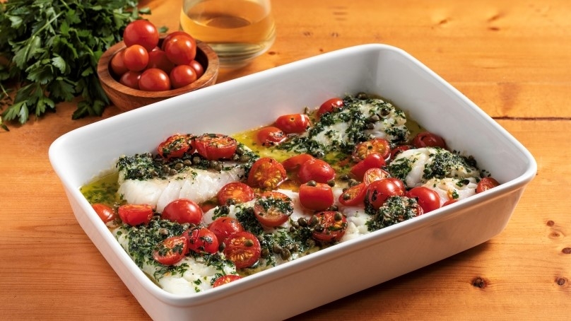 Herbed Butter Baked Cod in baking dish, cilantro, bowl of cherry tomatoes, bottle of oil, wood table