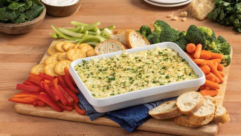 Hot Cheesy Spinach Dip in baking dosh, assorted cut veggies, cutting board, kitchen towel,  crackers, cut bread, bowl of spinach, bowl of sour cream, block of cheese, garnish, wood table