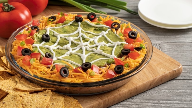 Large clear bowl of spooky layered spider dip, topped with diced tomatoes, black olives, shredded cheese, and sour cream, fresh tomatoes, scallions and white plates in background, gray wooden table