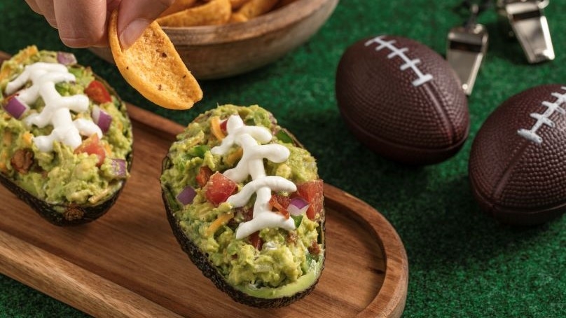 Loaded Guacamole Footballs on wooden serving tray, hand with corn chip, bowl of corn chips, mini footballs, whistles, green artificial grass table cloth