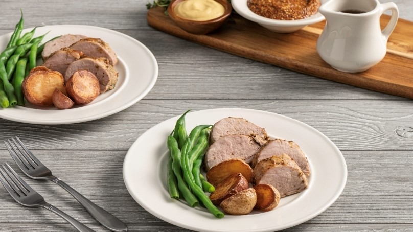 Two white plates with Maple-Glazed Roasted Pork Tenderloin and green beans. Gray wood table surface.. Wood tray with white syrup container, white bowl of brown sugar, dijon mustard.