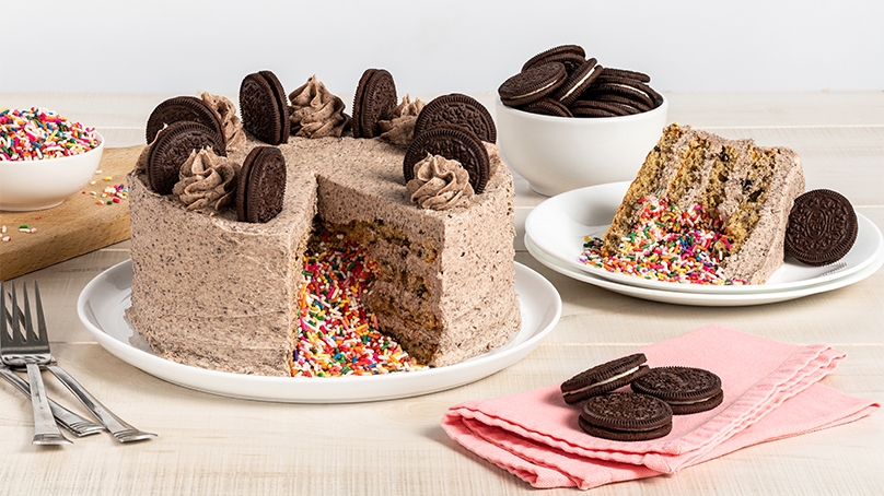 Oreo Birthday Cake, slice of cake on plate with oreo cookies, sprinkles spilling out of inside of cake, pink napkin with oreos, serving bowl of oreos in background with bowl of rainbow colored sprinkles, forks, wood table