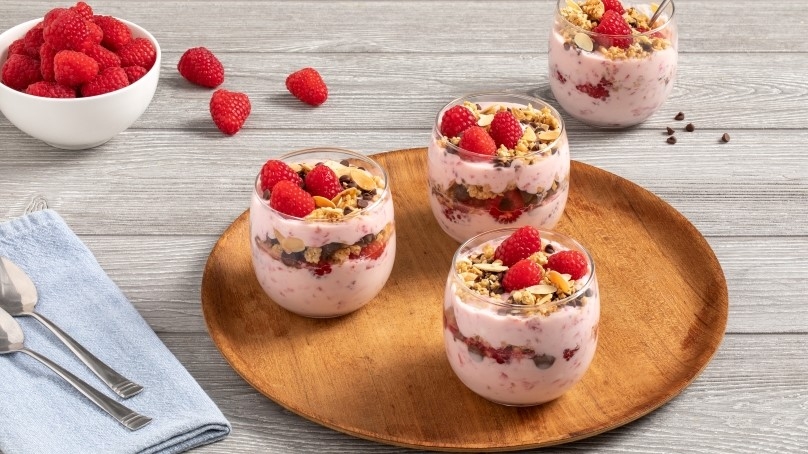 Raspberry Yogurt Parfaits in glasses on wooden serving tray, bowl of raspberries, napkin and spoons on gray wood table