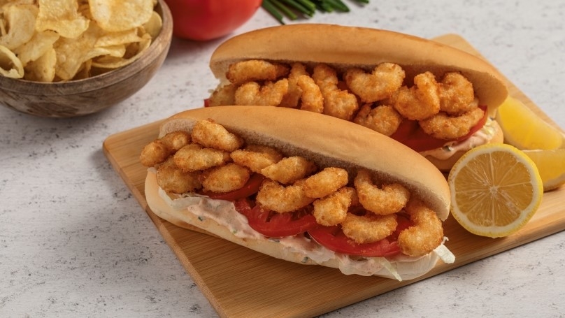 Shrimp Po Boy sandwiches on wooden cutting board with halved lemon, serving bowl of potato chips in background with fresh tomato and chives, white stone counter