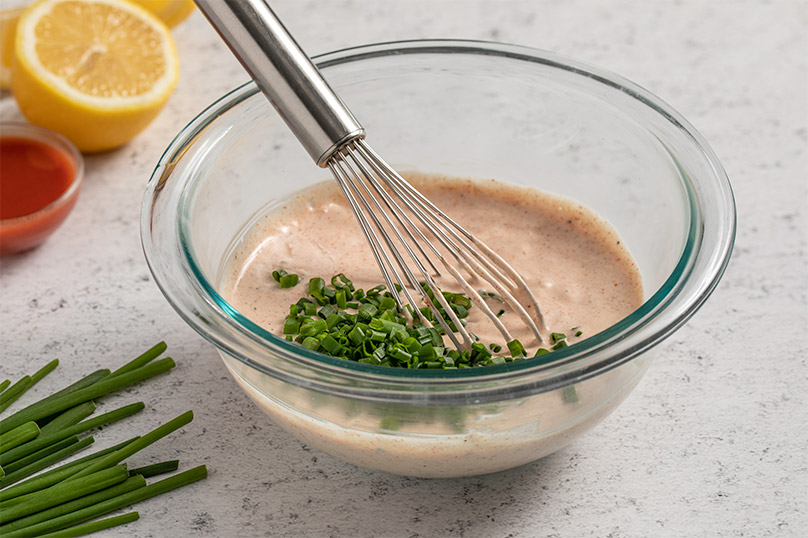 Whisk in a medium glass bowl mixing a remoulade sauce, fresh scallions for garnish, halved lemons in background, gray stone counter