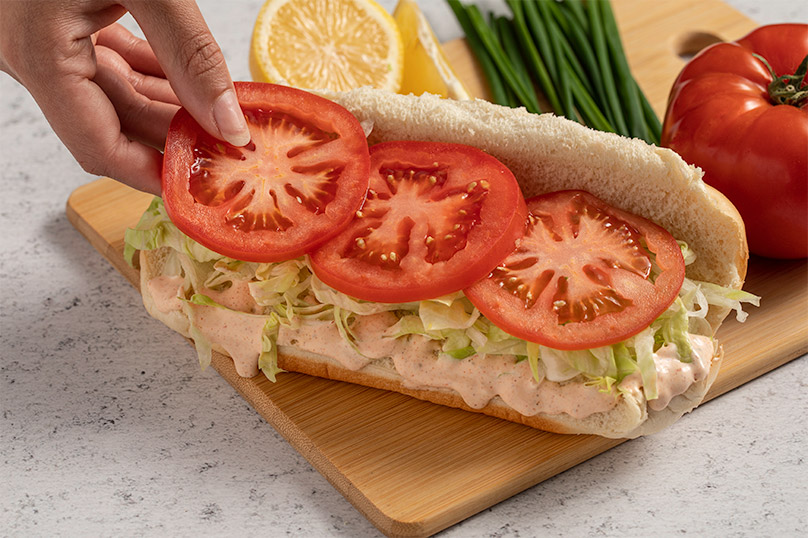 Hand adding tomatoes to Shrimp Po Boy sandwich on cutting board with scallions, fresh tomato, and halved lemon in background, white stone counter