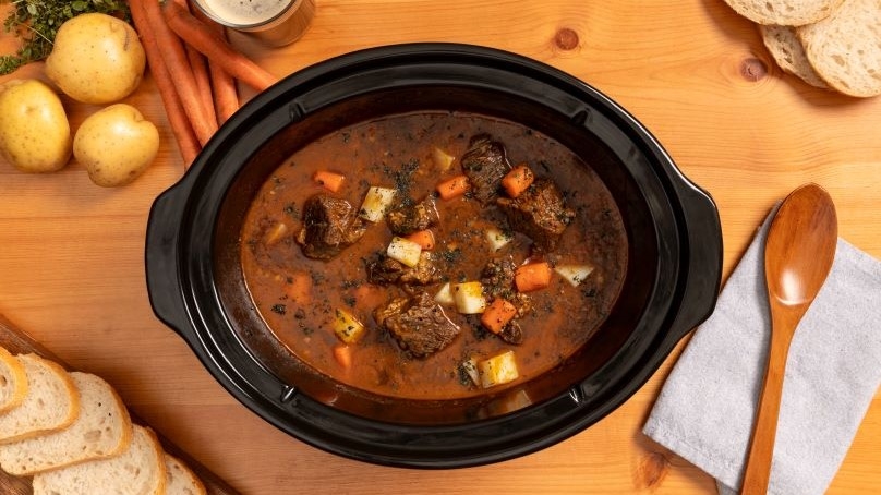 Slow Cooker filled with Irish Beef Stew, carrots, potatoes, slices of bread, wooden spoon on kitchen towel