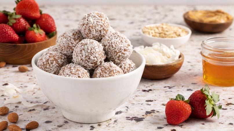 Bowl of strawberry breakfast bites, strawberries, bowl of coconut shavings, bowl of rolled oats, almonds, small jar of honey, stone counter