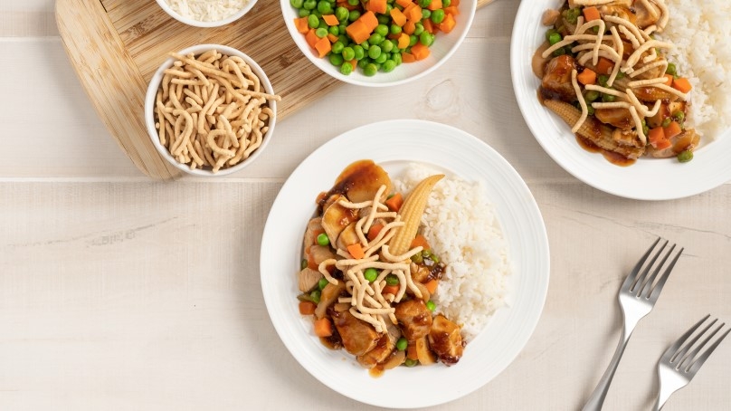 Two white plates with Teriyaki Chicken Stir Fry, two forks on the side, wood tray with bowl of Chow Mein noodles, bowl of carrots and peas, and bowl of rice