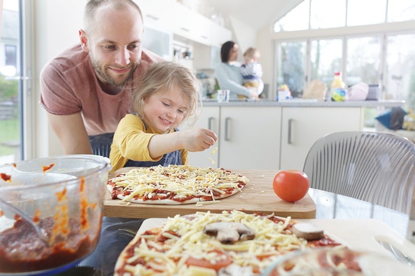 Father watching daughter spread cheese on homemade pizza, bright kitchen with family, mom and baby in background