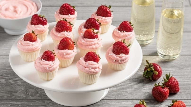 Serving dish of mini prosecco cupcakes garnished with fresh strawberries, glasses of prosecco in background with serving bowl of strawberry icing and fresh strawberries