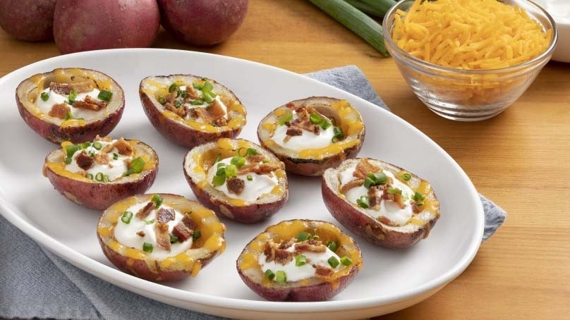 Loaded Mini Potato Skins on white serving dish, red potatoes, celery stalks, bowl of shredded cheddar cheese, cruet of sour cream, light blue napkin, wood table