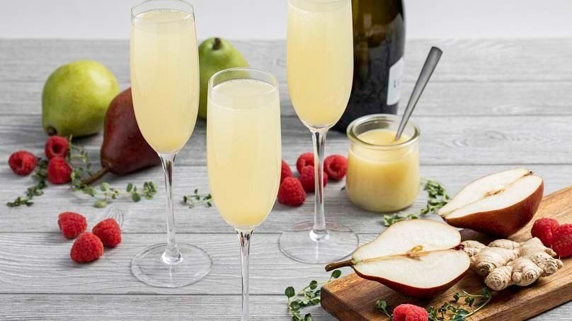 Sparkling Pear Mimosas in champaign flutes, sliced and whole pears, ginger, raspberries, honey jar, bottle of Proseco, wood table
