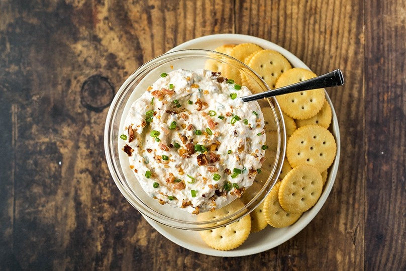 Million dollar dip in bowl with spoon on plate of crackers, wood table background