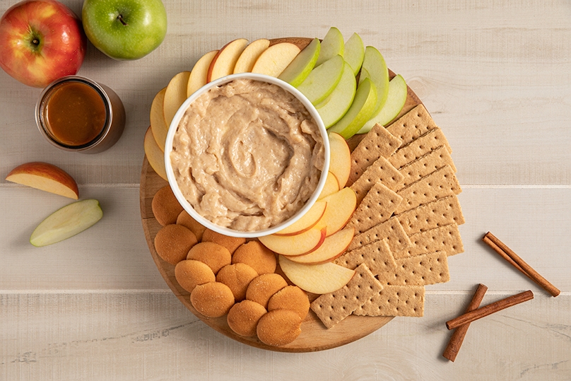 No Bake Apple Caramel Cheesecake Dip with fresh apple slices, graham crackers, and vanilla wafers on wooden board, serving dish of caramel, fresh apples and cinnamon sticks in background, light wood table