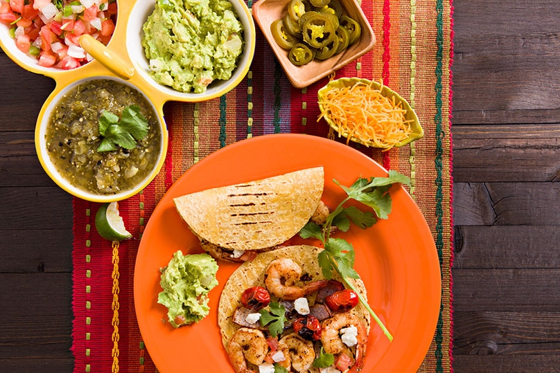 Shrimp Tacos on Plate, dishes of salse verde, pico de gallo, guacamole, sliced jalapenos and shredded cheese, mulit-colored striped woven table runner,  dark wood table