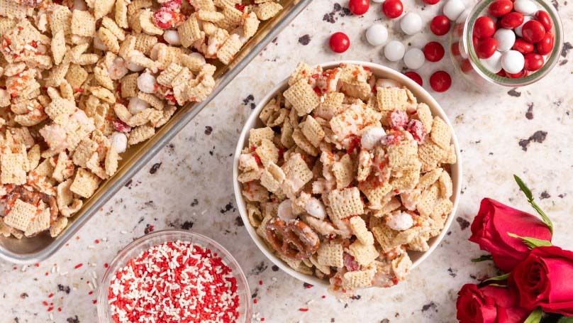 Sweet Valentine's Day Snack mix in a bowl and on a baking sheet, loose candies, bowl of sprinkles, roses, granite counter top