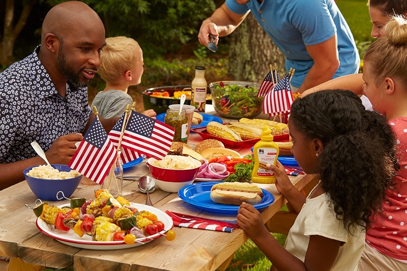 Family and friends gathered around picnic table eating hotdogs, kabobs, hamburgers, small USA flags, Food Lion mustard