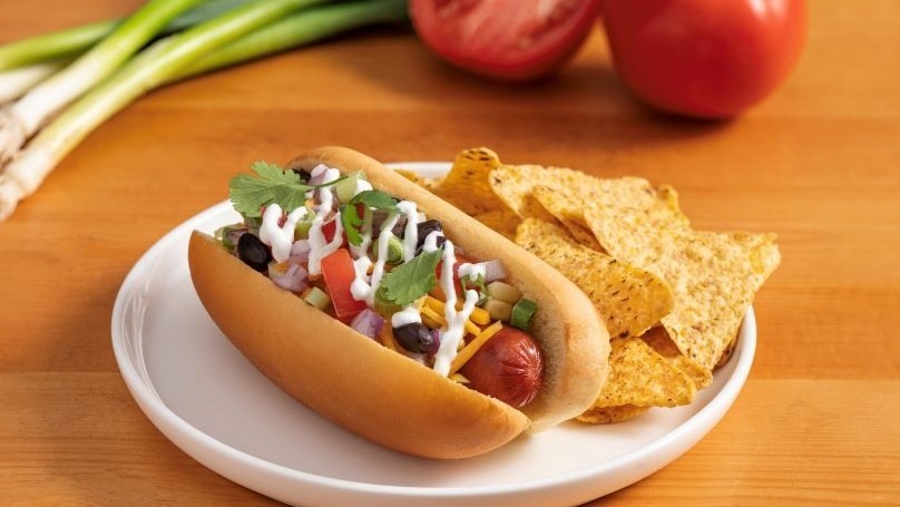 Tex-Mex Hot Dog with tortilla chips on white plate, fresh tomato and green onion in background, light wood table