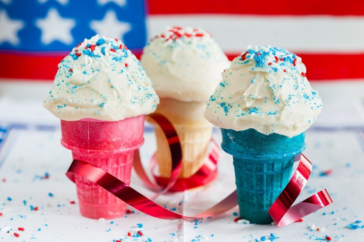 Red white and blue ice cream cone cupcakes, red white and blue sprinkles, red ribbon around ice cream cones, USA flag in background