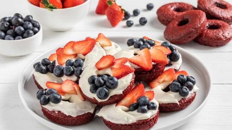 Red white and blue cake donuts topped with blueberries and strawberry slices on white plate, fresh strawberries and blueberries in serving bowls, cake donuts, white wood table