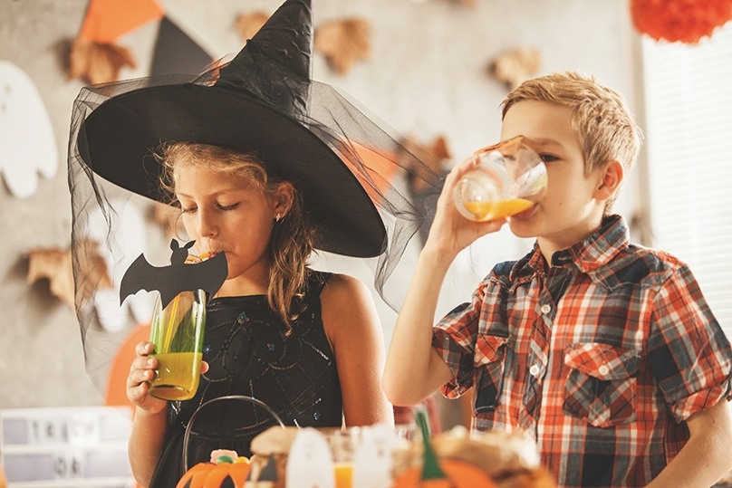 Boy and a girl standing by the table with treats at the Halloween party. They are drinking juice.