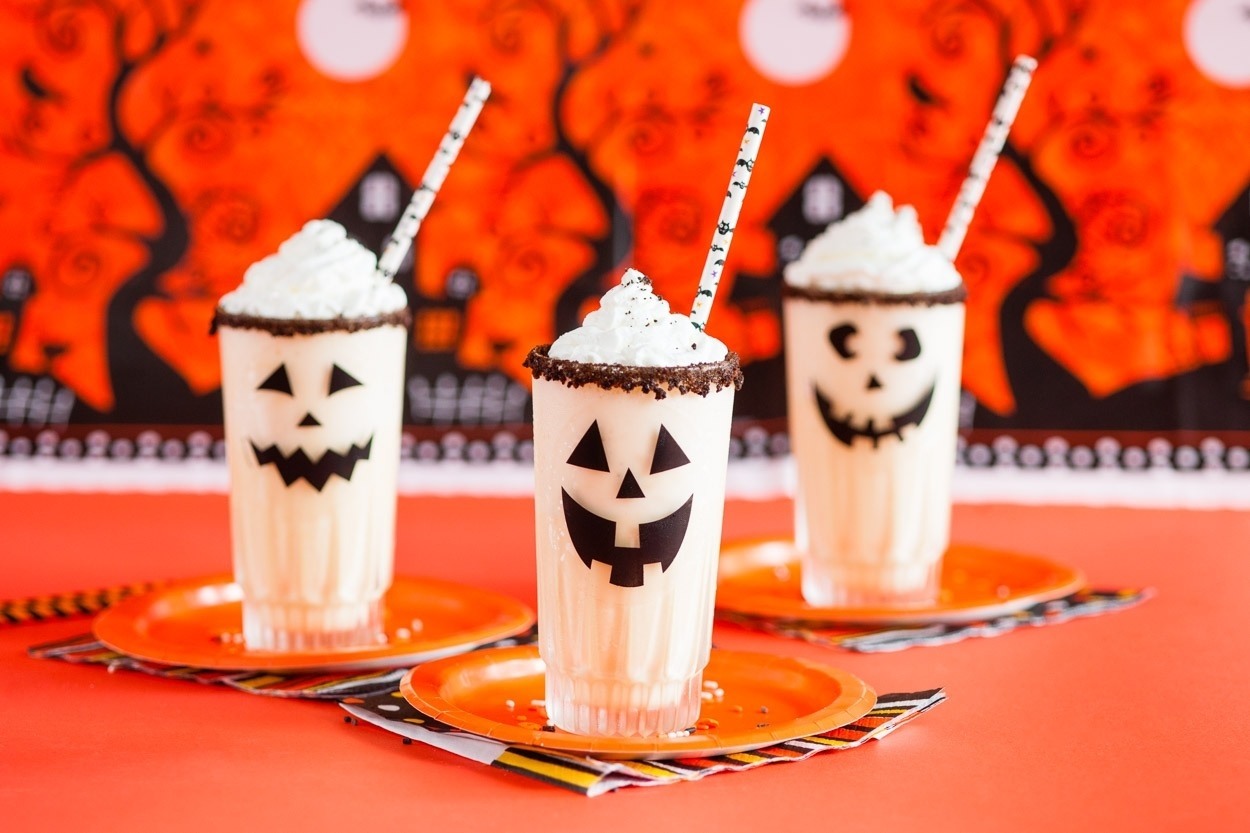 Vanilla milkshakes with jack-o-lantern faces on the outside, halloween themed decorations in the background