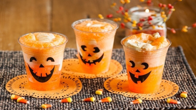 3 Orange sodas in plastic cups with jack-o-lantern faces on the outside, candy corn on table, gray placemat on wooden table