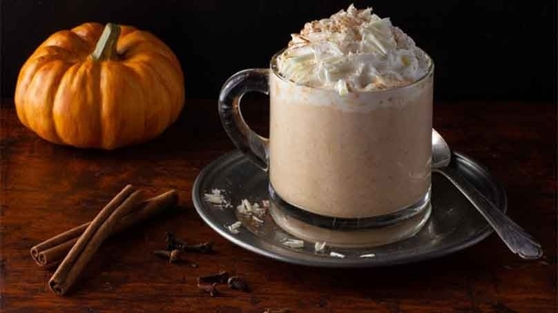 Cup of white hot chocolate topped with whipped cream on plate with spoon, fresh stick of cinnamon and small pumpkin in background, wood table