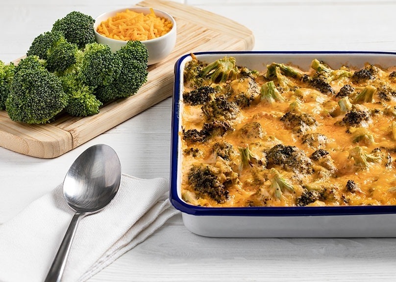 Keto Broccoli &amp; Cheddar Casserole in baking pan, cutting board, broccoli flourettes, cheese in cruet, spoon, napkin,, white counter top