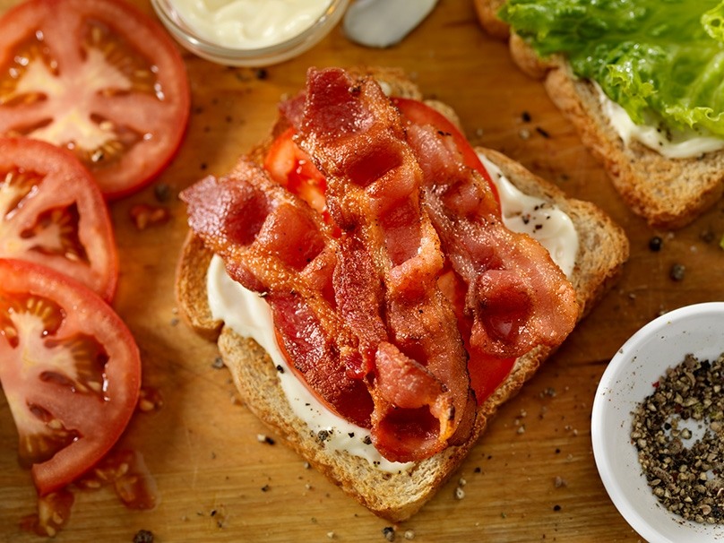 Closeup of bacon on top of bread with mayonnaise and lettuce, fresh tomato slices, serving bowl of pepper, serving dish of mayonnaise, wood table