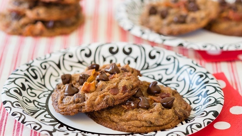 Chocolate chip cookies topped with bacon crumbles on paper plates, on table with red striped tablecloth