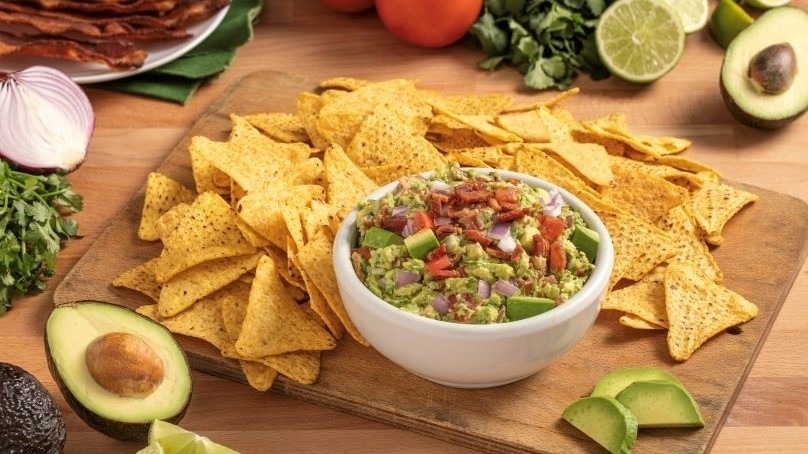 Serving bowl of bacon guacamole with fresh tortilla chips on wooden cutting board, fresh avocado, onion, tomato, bacon strips, halved limes, surrounding cutting board