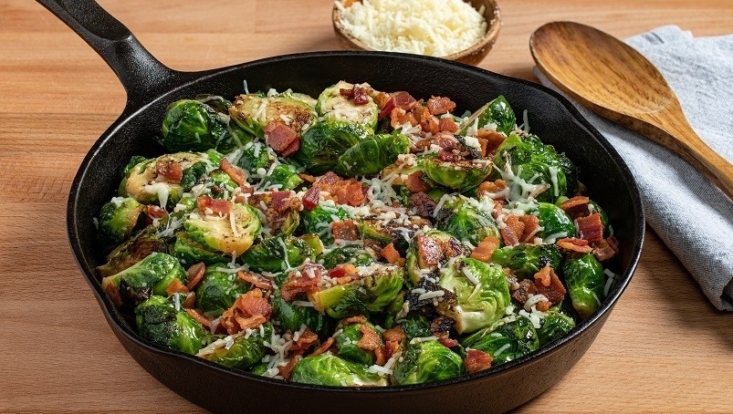 Keto Brussel sprouts with bacon in iron pan with serving bowl of shredded cheese, wooden spoon, blue kitchen towel, wood table